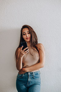 Young woman using phone while standing against wall