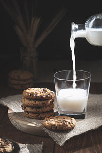 Chocolate chip cookies and milk bottle spilling milk in glass on wooden base, dark background. 