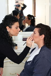 Side view of beautician applying make-up on young woman while sitting in front of mirror
