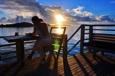 Man sitting on bench by sea against sky during sunset