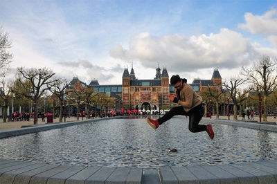 Young man jumping on retaining wall by pond in front of rijksmuseum