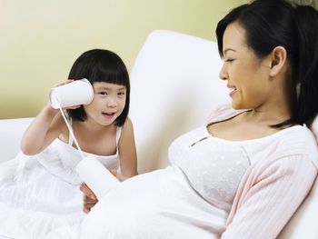 Girl listening pregnant mother abdomen with tin can phone on sofa