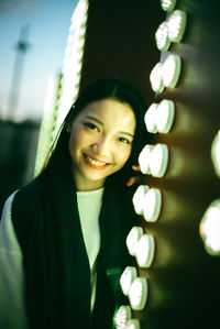 Portrait of smiling young woman standing by illuminated wall