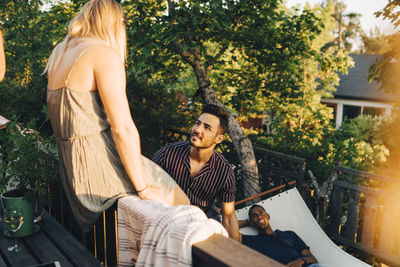 Friends talking at railing while man resting in hammock at yard