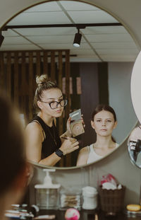 A young makeup artist looks in the mirror at a girl doing makeup.