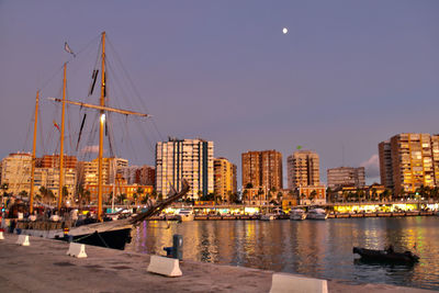 Sailboats in river by buildings against clear sky at dusk