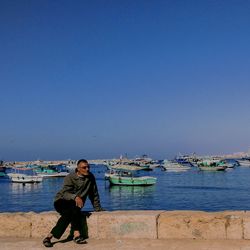 Mature man sitting on retaining wall by sea against clear blue sky