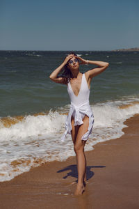 Woman in a white swimsuit and a straw hat stands on an empty sandy beach