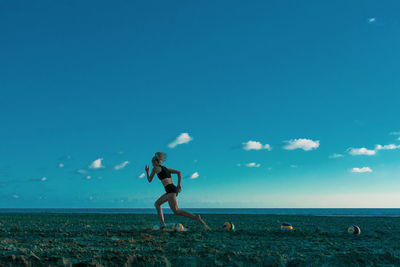 Side view of young woman running while exercising at beach against blue sky