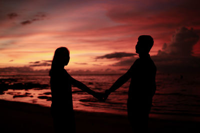 Silhouette couple holding hands while standing on beach during sunset