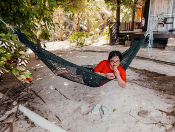 Happy asian teenager in casual outfit relaxing in hammock while using mobile phone. social media.