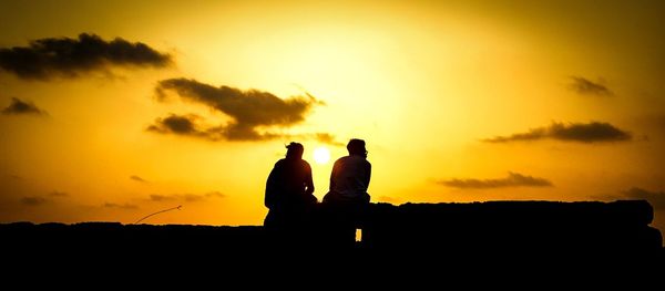 Low angle view of silhouette friends sitting on retaining wall against orange sky during sunset