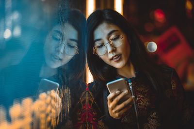 Smiling young woman using smart phone by window with reflection at night 