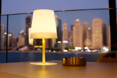 A burning lamp and ashtray stand on a table in a cafe with a night view of marina dubai, close-up