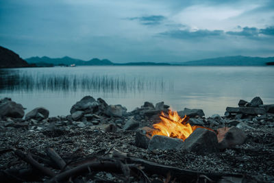 Bonfire at lakeshore against sky during sunset