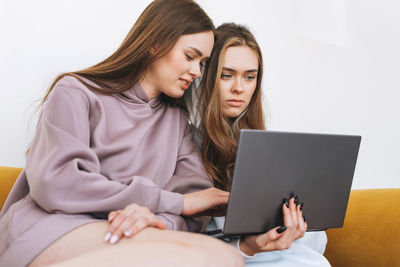 Young women sisters twins teenagers in hoodie beautiful girls using laptop sitting on yellow couch 