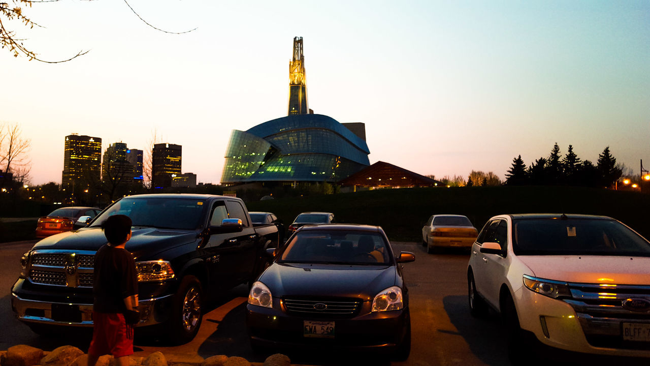 car, land vehicle, transportation, mode of transport, building exterior, architecture, city, built structure, street, clear sky, traffic, sunset, road, city life, travel, city street, tall - high, tree, tower, on the move