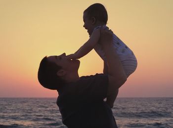 Playful mother carrying baby at beach against clear sky during sunset