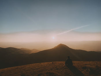 Silhouette woman sitting on grass against mountain and sky during sunset