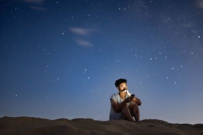 Side view of young man sitting on land against sky at night