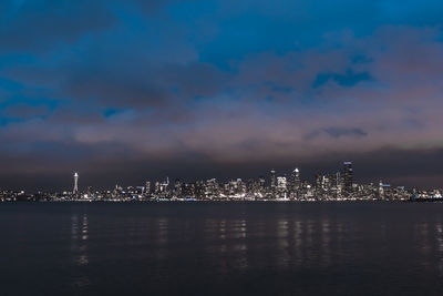 Illuminated city at waterfront against cloudy sky