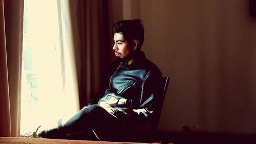 Thoughtful young man looking through window while sitting on chair at home
