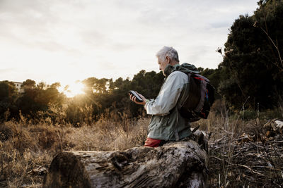 Senior male hiker using mobile phone sitting on wooden log during weekend