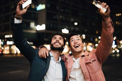 Happy man and male friend looking up with smart phones while gesturing in city at night