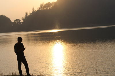 Silhouette man standing at lakeshore against trees during sunset