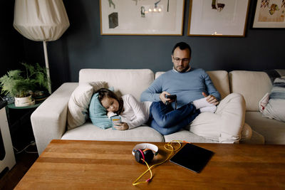 Father and daughter using mobile phones on couch in living room at home