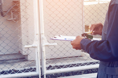 Midsection of man holding clipboard against chainlink fence