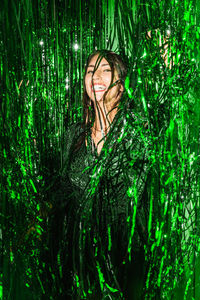 Delighted female wearing festive dress playing with green shiny tinsel stripes at party while looking at camera