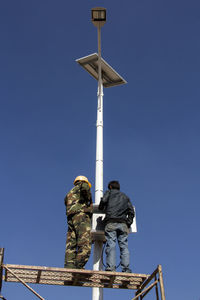 Rear view of men standing on solar powered street light against clear blue sky