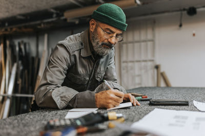 Bearded male entrepreneur wearing eyeglasses while writing at workbench