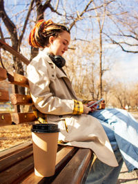 Coffee in a cup stands on a bench in the park and a young modern woman in a raincoat sits on a bench