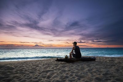 Side view of young man sitting on log at beach against sky during sunset