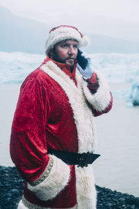Man in santa costume talking on smart phone while standing by lagoon during winter