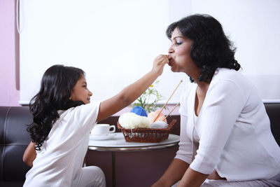 Side view of smiling daughter feeding food to pregnant mother in living room at home