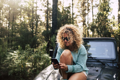 Woman using mobile phone while sitting on vehicle in forest