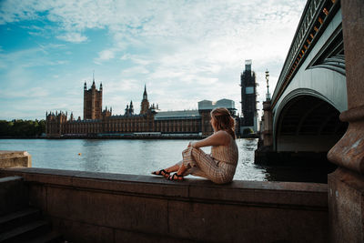 Woman sitting on bridge over river in city against sky