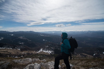 Side view of man with backpack standing on mountain against cloudy sky