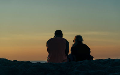 Rear view of couple sitting on rock at beach against sky