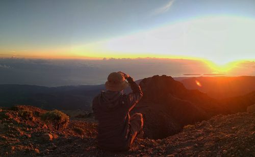 Rear view of person sitting on cliff at mount rinjani against sky during sunset