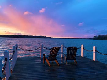 Wooden chairs by sea against sky during sunset