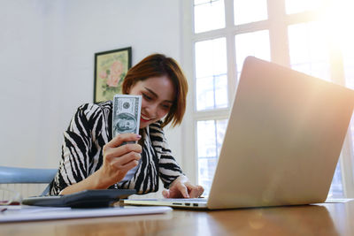 Smiling businesswoman using laptop while holding money at office desk