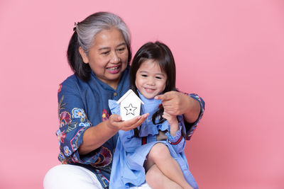 Smiling grandmother showing model home to granddaughter against pink background