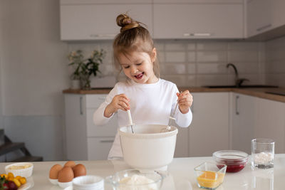 Smiling baby girl 3-4 year old making dough in white bowl for cake on kitchen table at home close up