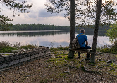 Rear view of man sitting by lake against trees