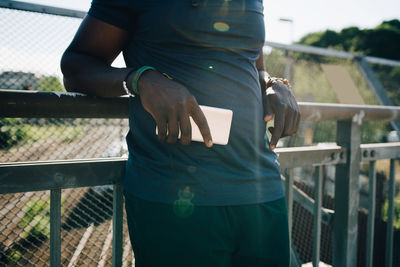 Midsection of sportsman holding mobile phone while leaning on railing during sunny day