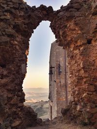 Bells hanging on building seen through abandoned stone wall during sunset